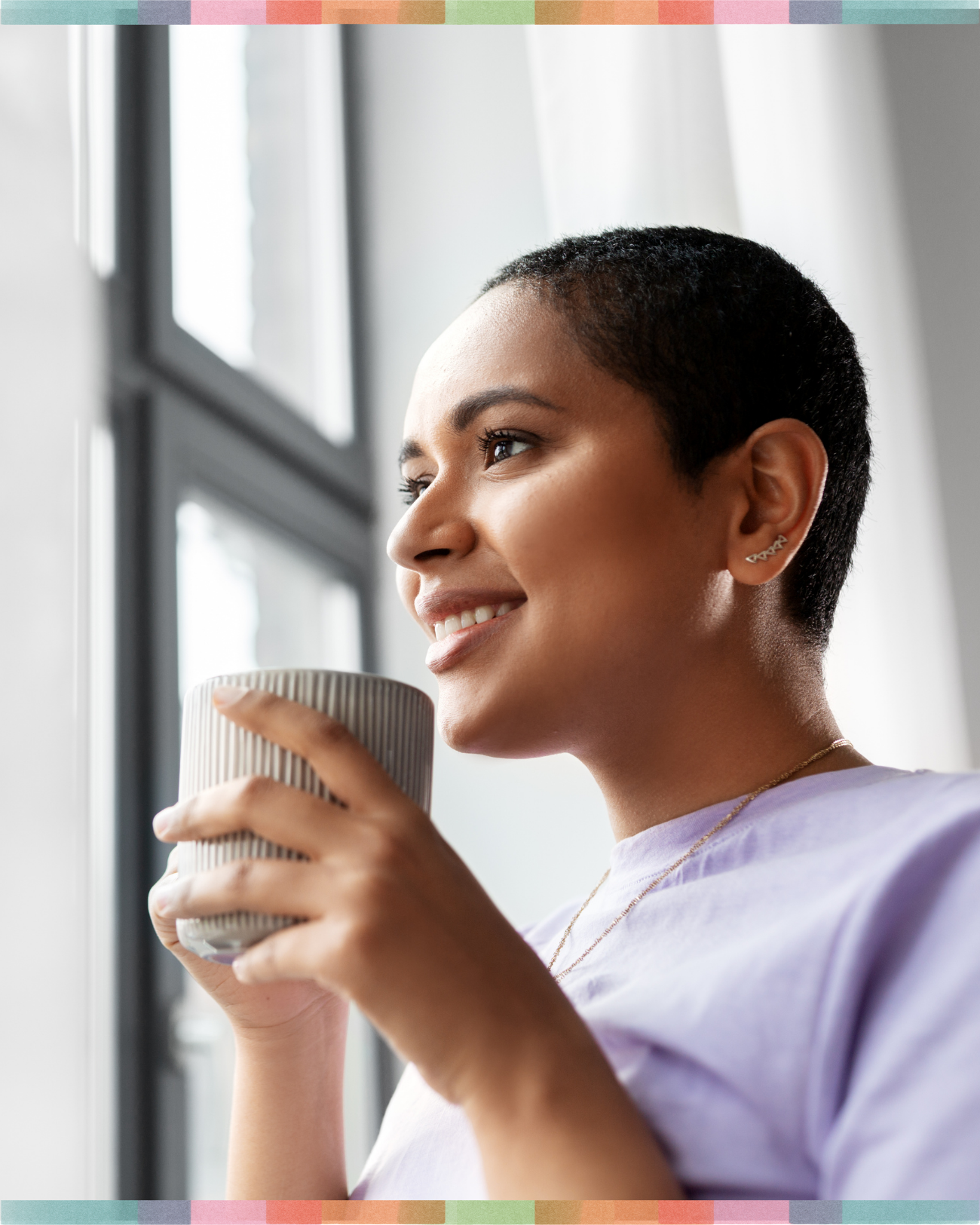 Woman with short curly hair smiling while holding a beige textured ceramic mug near a window with natural light.