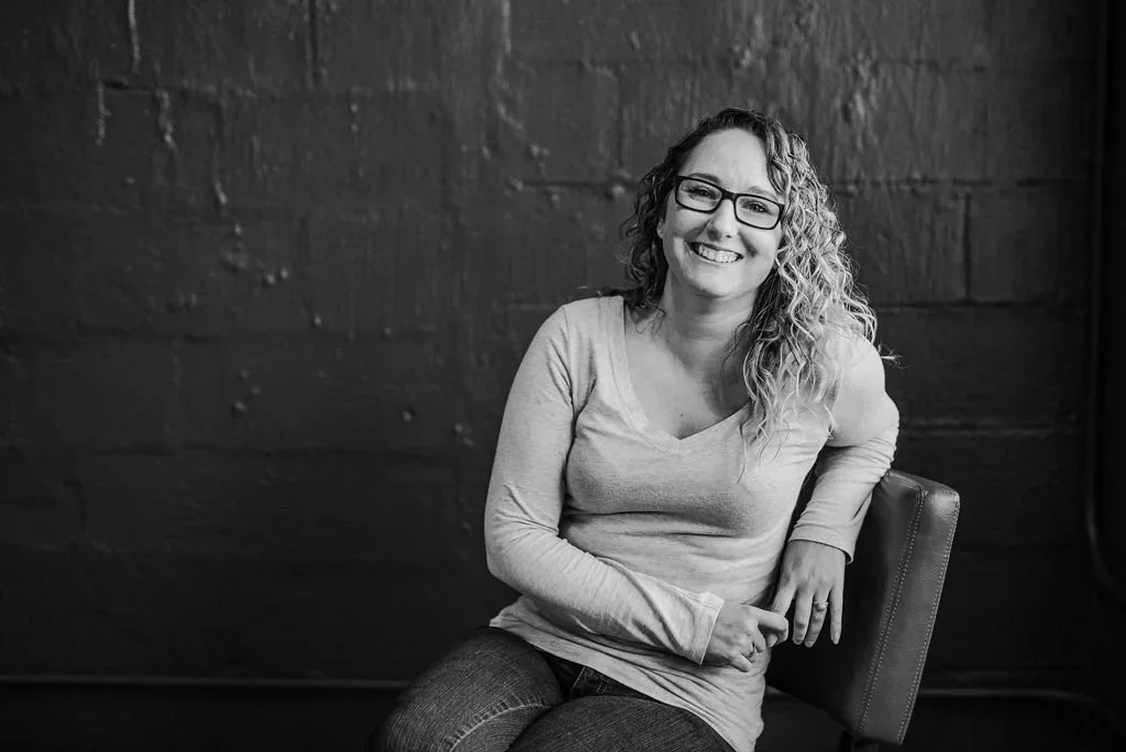 Black-and-white portrait of a smiling woman with curly hair and glasses seated against a dark textured wall