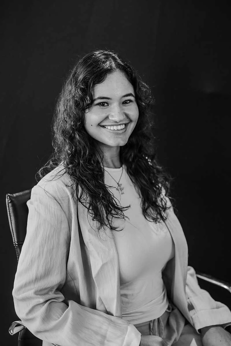 Black-and-white portrait of a smiling woman with long curly hair seated against a dark background.