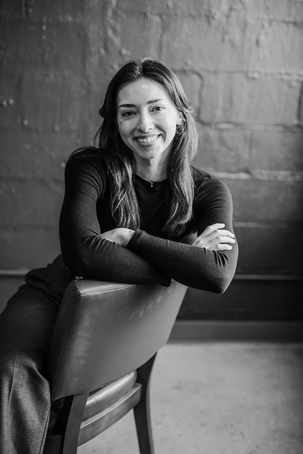 Black-and-white portrait of a smiling woman with crossed arms leaning over a chair in front of a textured wall