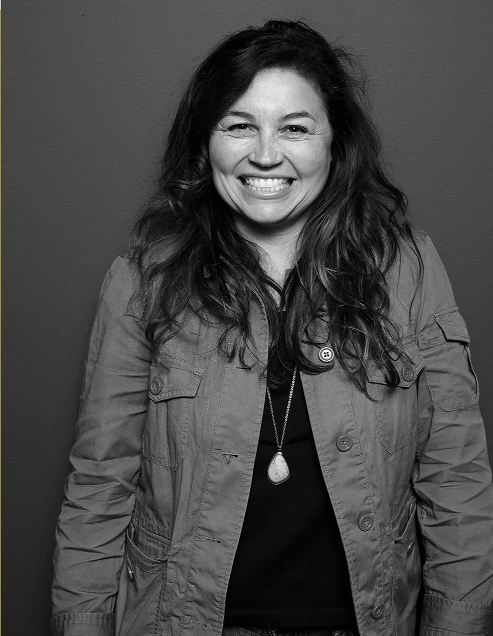Black-and-white portrait of a smiling woman with long hair wearing a utility jacket and pendant necklace