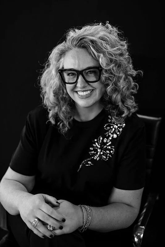 Black-and-white portrait of a smiling curly-haired woman wearing glasses, a black top with a jeweled floral design, rings, and bracelets.