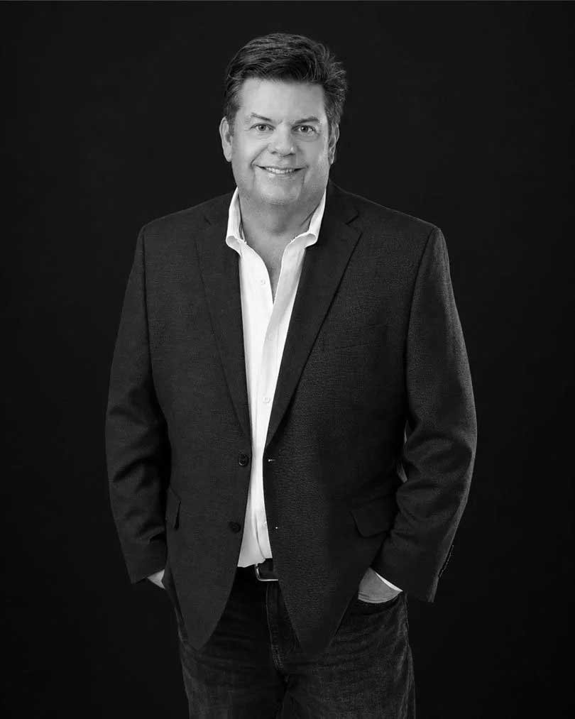 Black-and-white portrait of a smiling man in a blazer and open-collar shirt against a dark background