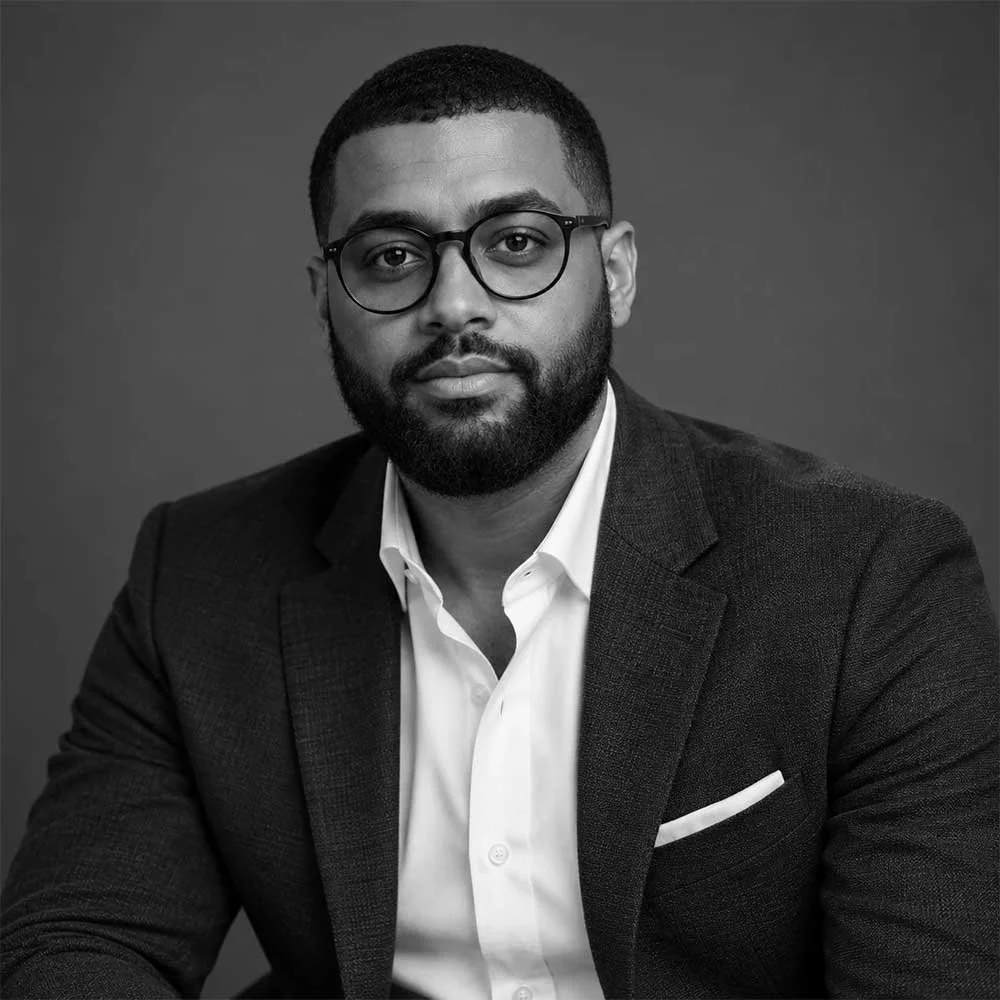 Black-and-white professional headshot of a bearded man wearing round glasses, a suit jacket, and white shirt