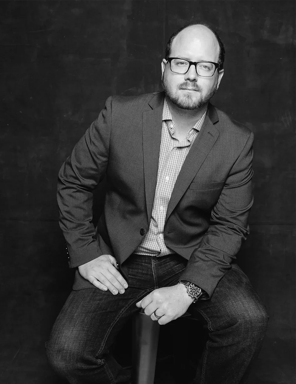 Black-and-white portrait of a bald, bearded man wearing glasses, a blazer, checked shirt, jeans, and a wristwatch.