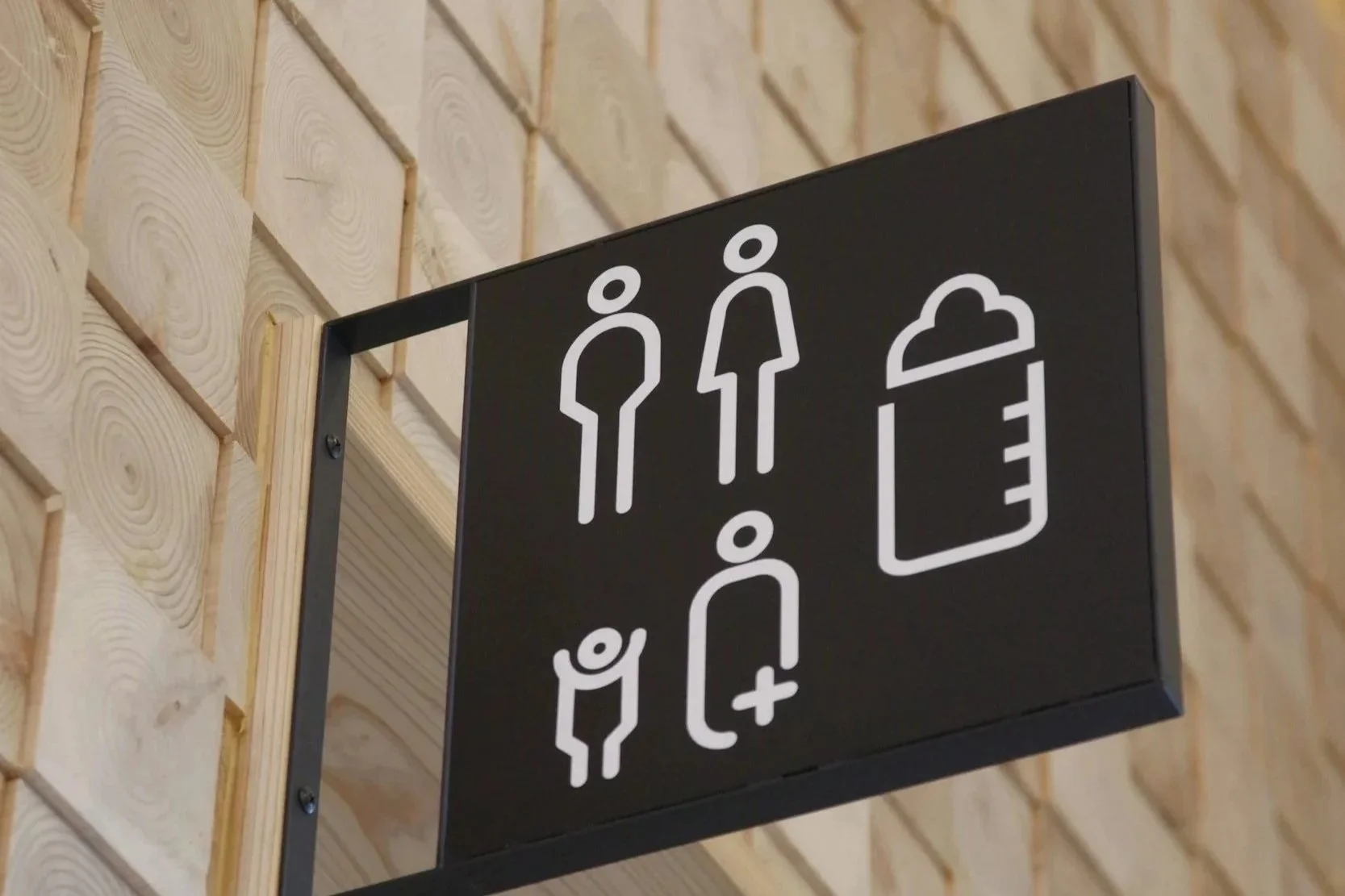 A black sign with white icons indicating a men's bathroom, a women's bathroom, a unisex bathroom, and a baby changing station, mounted on a wooden wall.