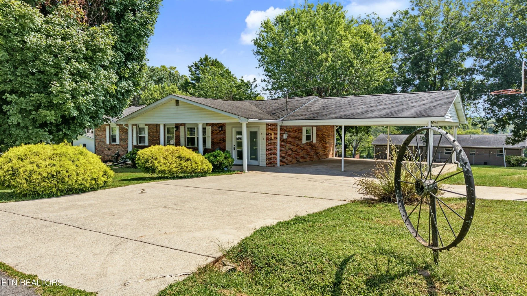 A single-story brick house with a front porch, surrounded by green bushes and trees. The driveway is concrete, and there is a vintage metal wagon wheel ornament on the lawn. The background features a clear blue sky with a few clouds and neighboring houses.
