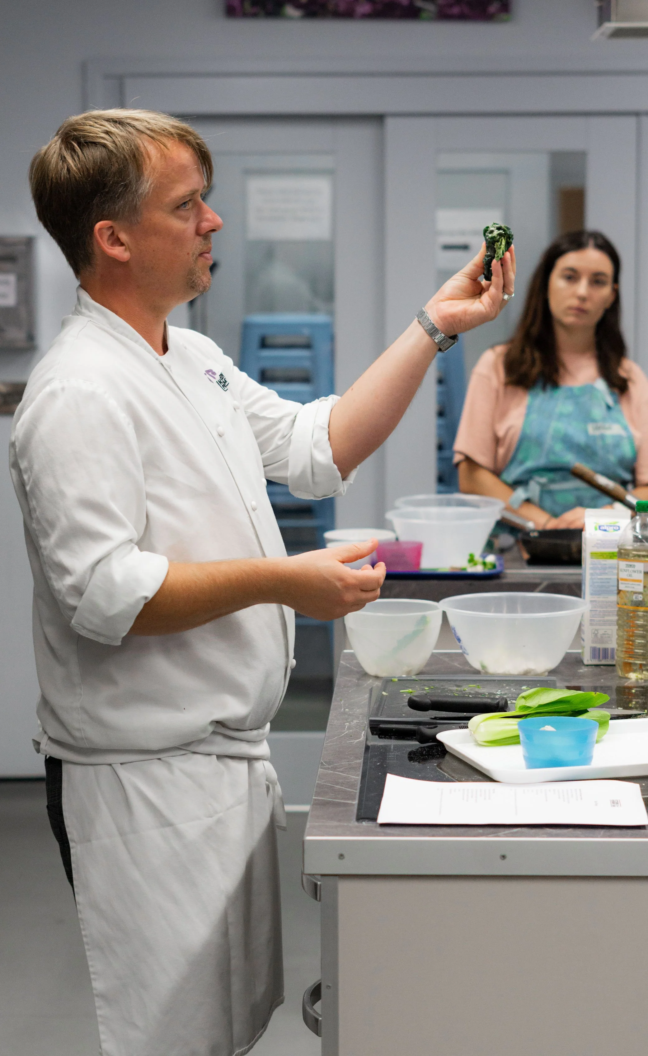 A chef holding a piece of food and a woman watching in a commercial kitchen with bowls, utensils, and ingredients on the counter.