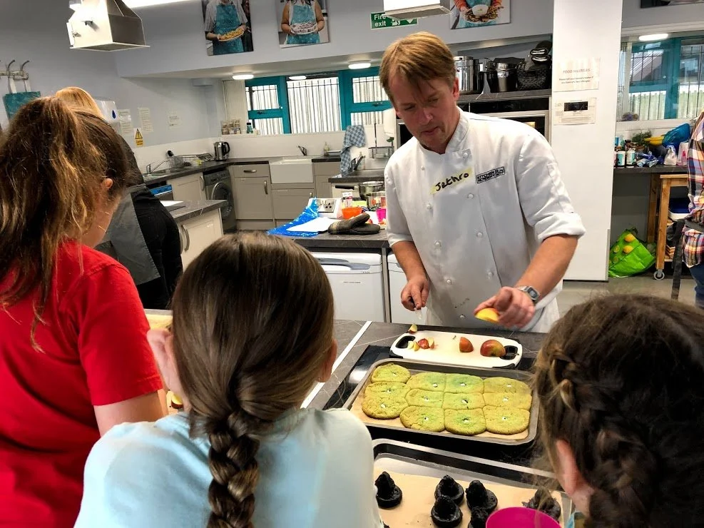 A chef demonstrating baking to a group of children in a commercial kitchen.