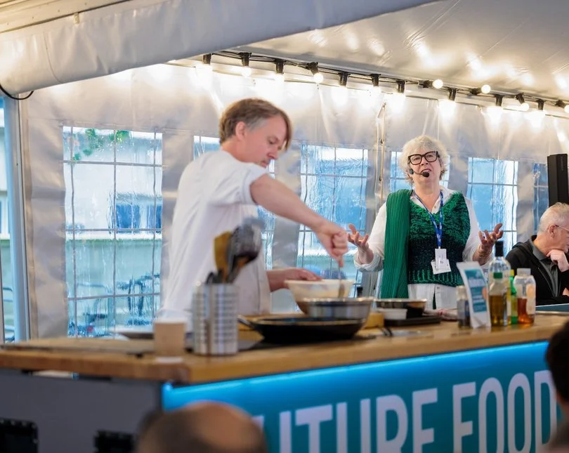 A woman chef is working at a cooking demonstration station, stirring a bowl, while an older woman with glasses and a green scarf gesturing and speaking in front of an audience inside a tent.
