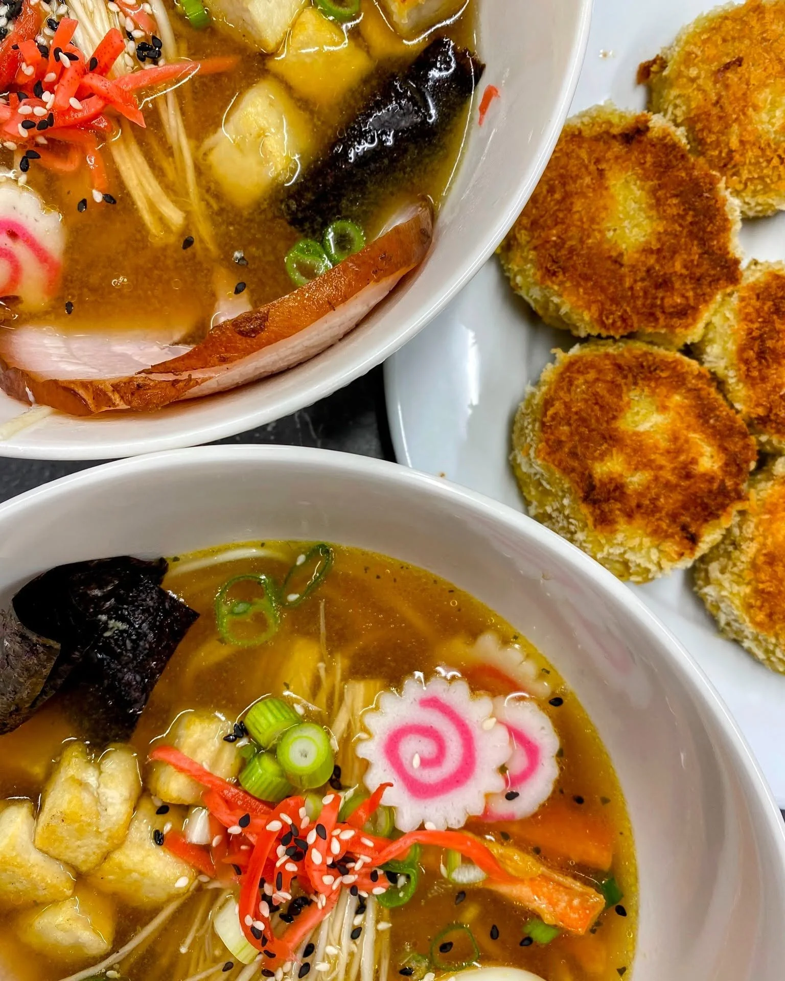 Close-up of bowls of ramen with toppings including narutomaki fish cake, green onions, and black sesame seeds, and a plate of breaded fried food on the side.