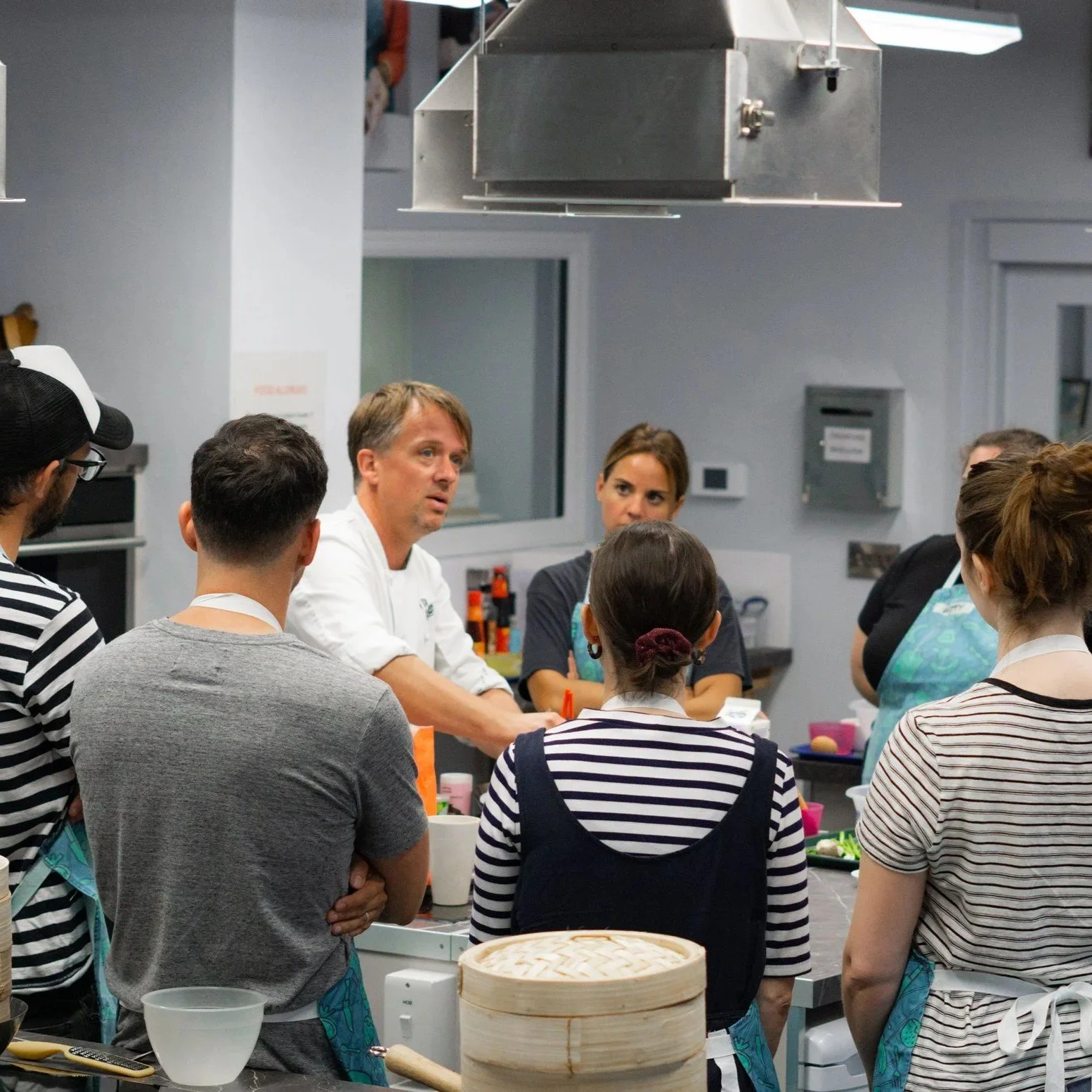Group of people gathered around a chef in a kitchen, listening attentively. The chef, wearing a white coat, appears to be explaining or demonstrating something. The kitchen is equipped with various tools and ingredients, including a bamboo steamer and aprons.