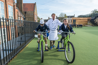 A chef demonstrating smoothie bikes with two children in a schoolyard or sports field.