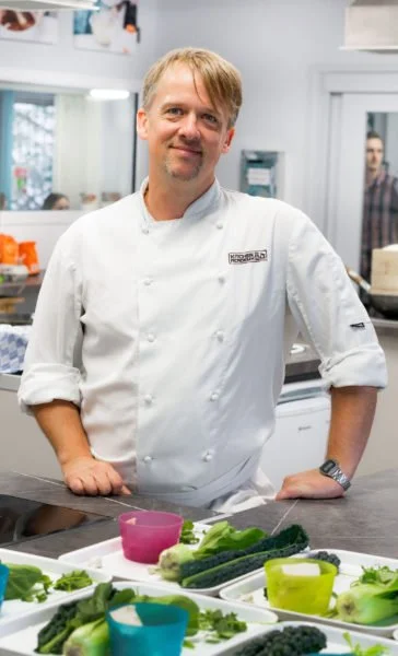 A male chef standing in a kitchen, surrounded by vegetables on a counter.