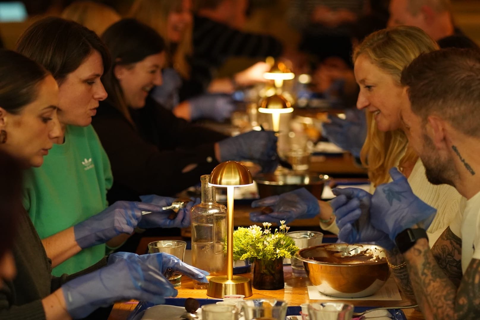 People sitting at a long table engaging in a group cookery activity, wearing gloves, with warm lighting and small lamps on the table.