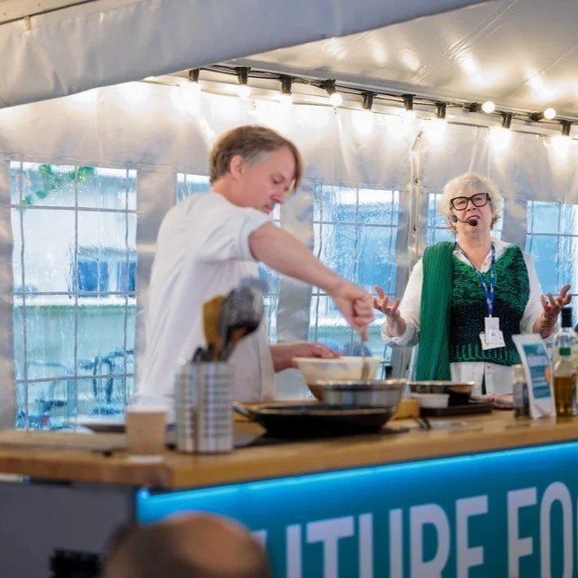 Chef and woman with microphone speaking at a cooking demonstration inside a tent at an outdoor event.