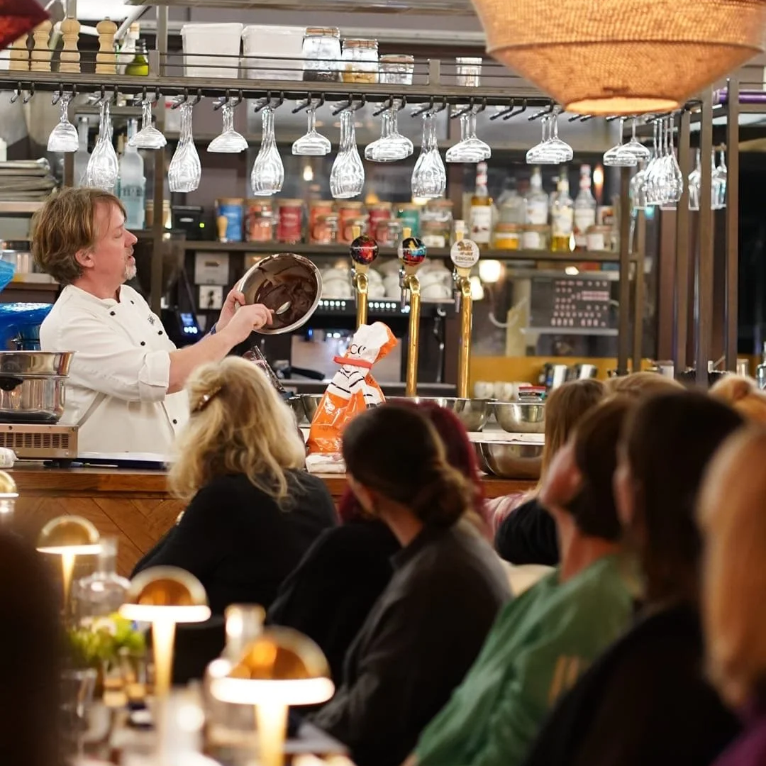 A chef giving a cooking demonstration at a restaurant bar, with guests watching attentively.