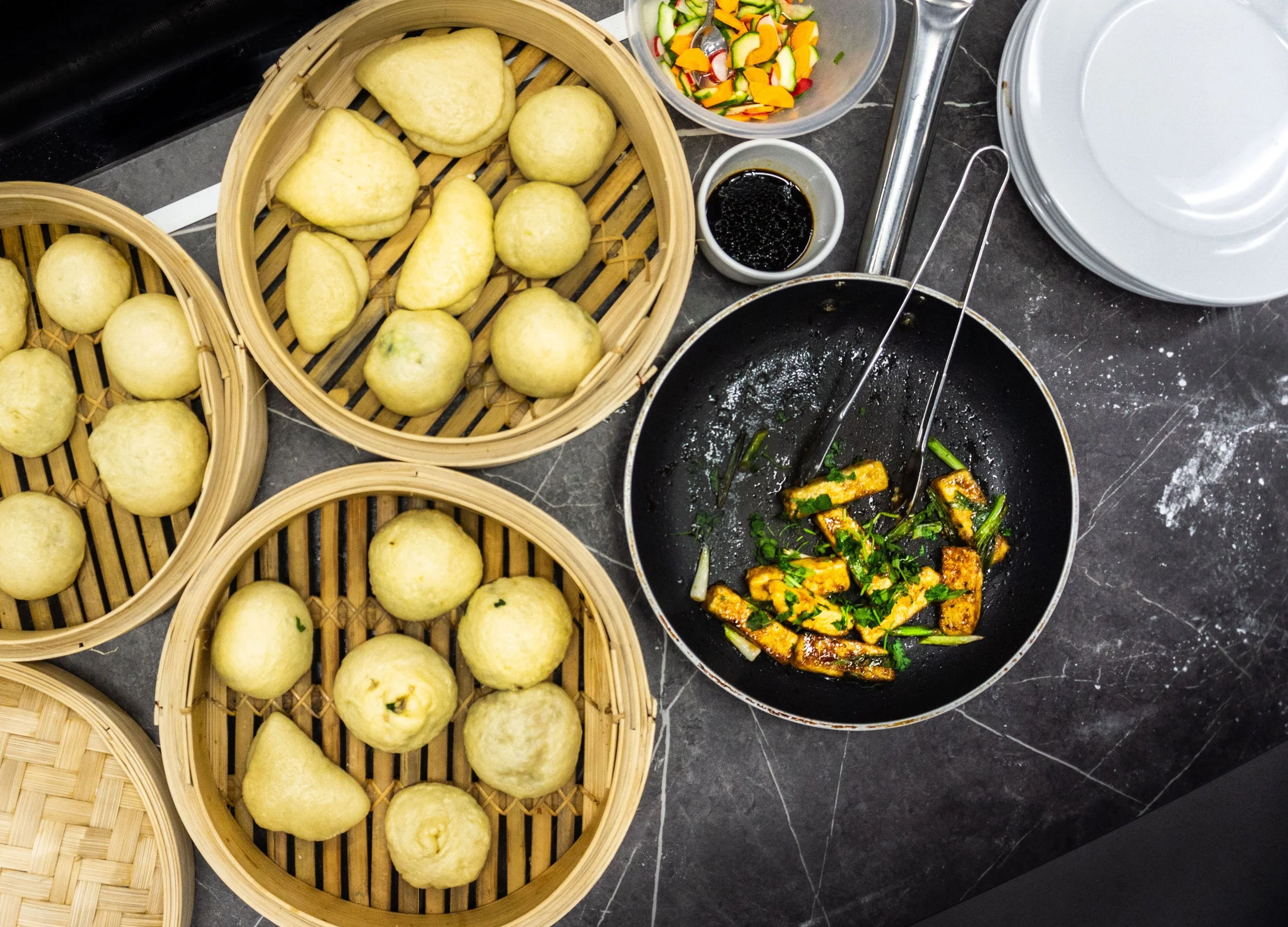 A kitchen countertop with three bamboo steamers filled with steamed buns, a bowl of mixed chopped vegetables, a small dish of soy sauce, and a frying pan with grilled vegetables and green herbs.