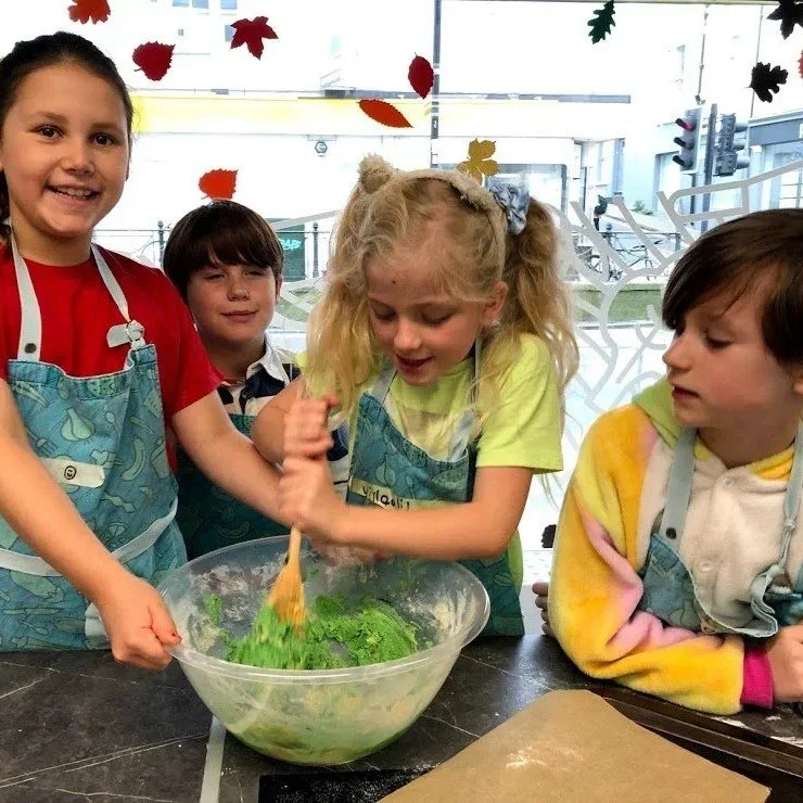 Four children in aprons making a salad or dough, with colorful autumn leaves hanging in the background.