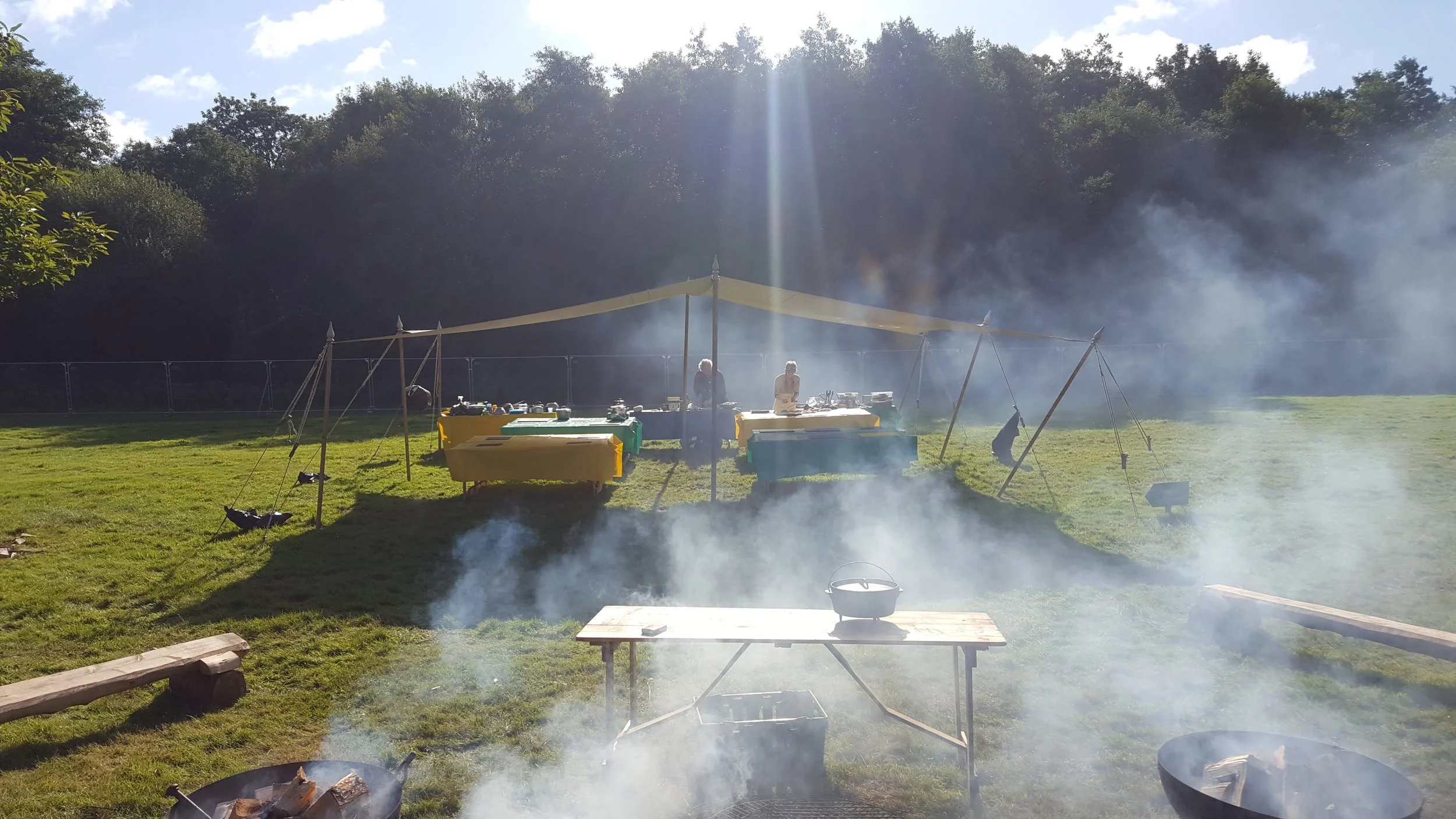 Outdoor cookery over fire with a table set up under a beige canopy in a grassy area, smoke rising from a fire pit, and trees in the background.