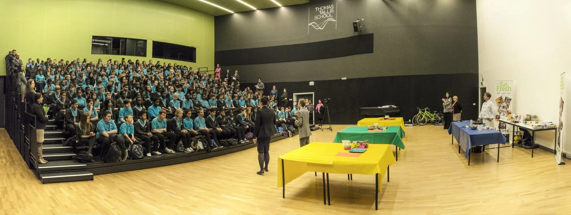 Students in blue and black uniforms seated in an auditorium while chef performs a cookery demonstration as part of a school assembly.