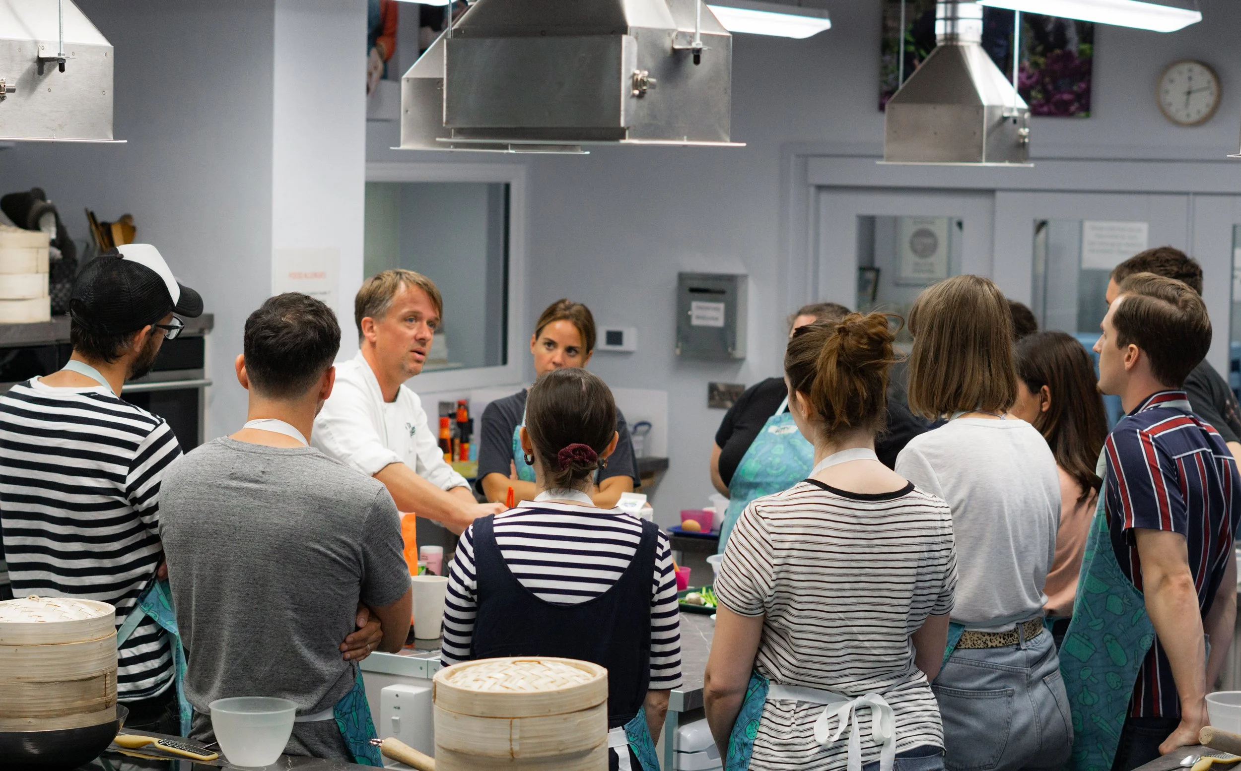 A group of people gathered around a cooking demonstration in a kitchen. The chef is talking, and the attendees are listening attentively. The kitchen has stainless steel appliances and hanging exhaust hoods.