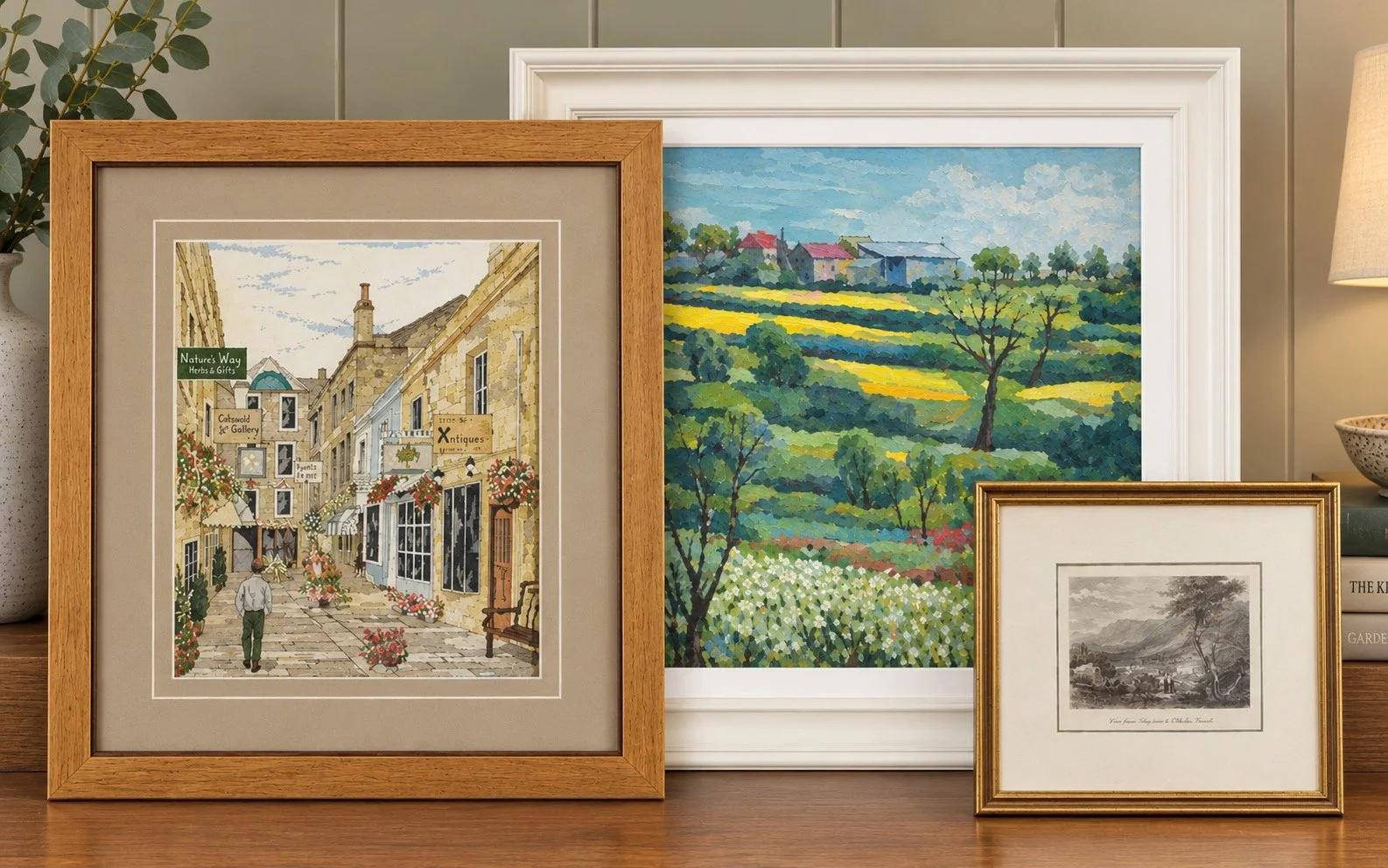 Three framed artworks displayed on a wooden surface: a colourful embroidery of a village street scene, a vibrant oil painting of a rural landscape, and a vintage print of mountains and trees.