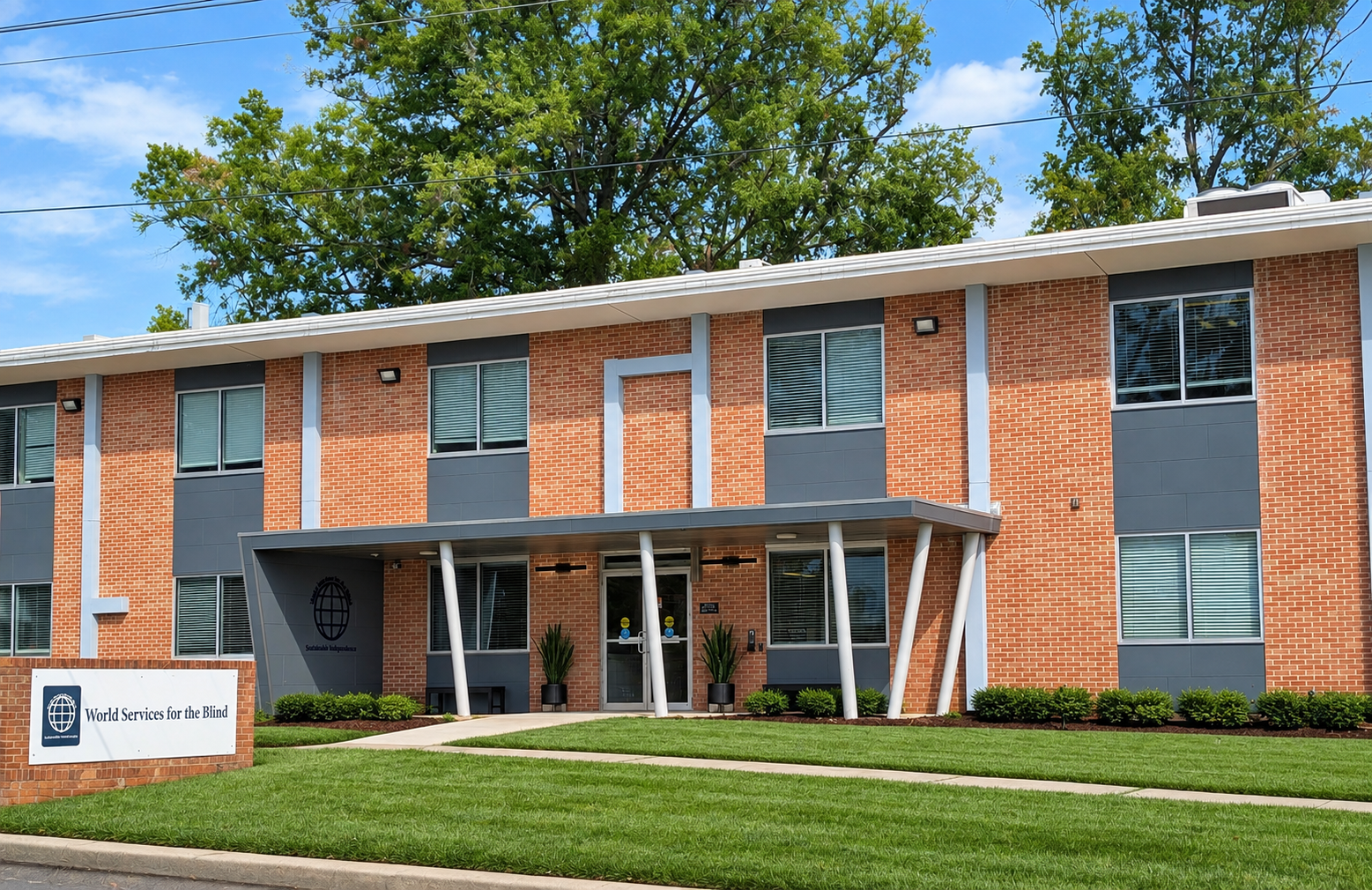 Building with a sign that reads 'World Services for the Blind', covered entrance, large windows, and landscaped lawn with green grass and bushes, trees in the background under a blue sky.