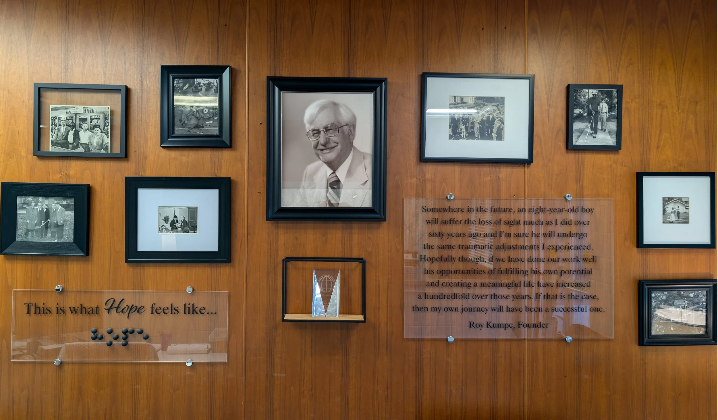 A wooden wall decorated with framed black and white photographs and a quote about hope and life's journey. The photos include people in various settings, such as a group of individuals, a person with a saw, and a person with a walking stick. There is also a small glass award or trophy and a clear acrylic plaque with a quote from Roy Kumpe, the founder.