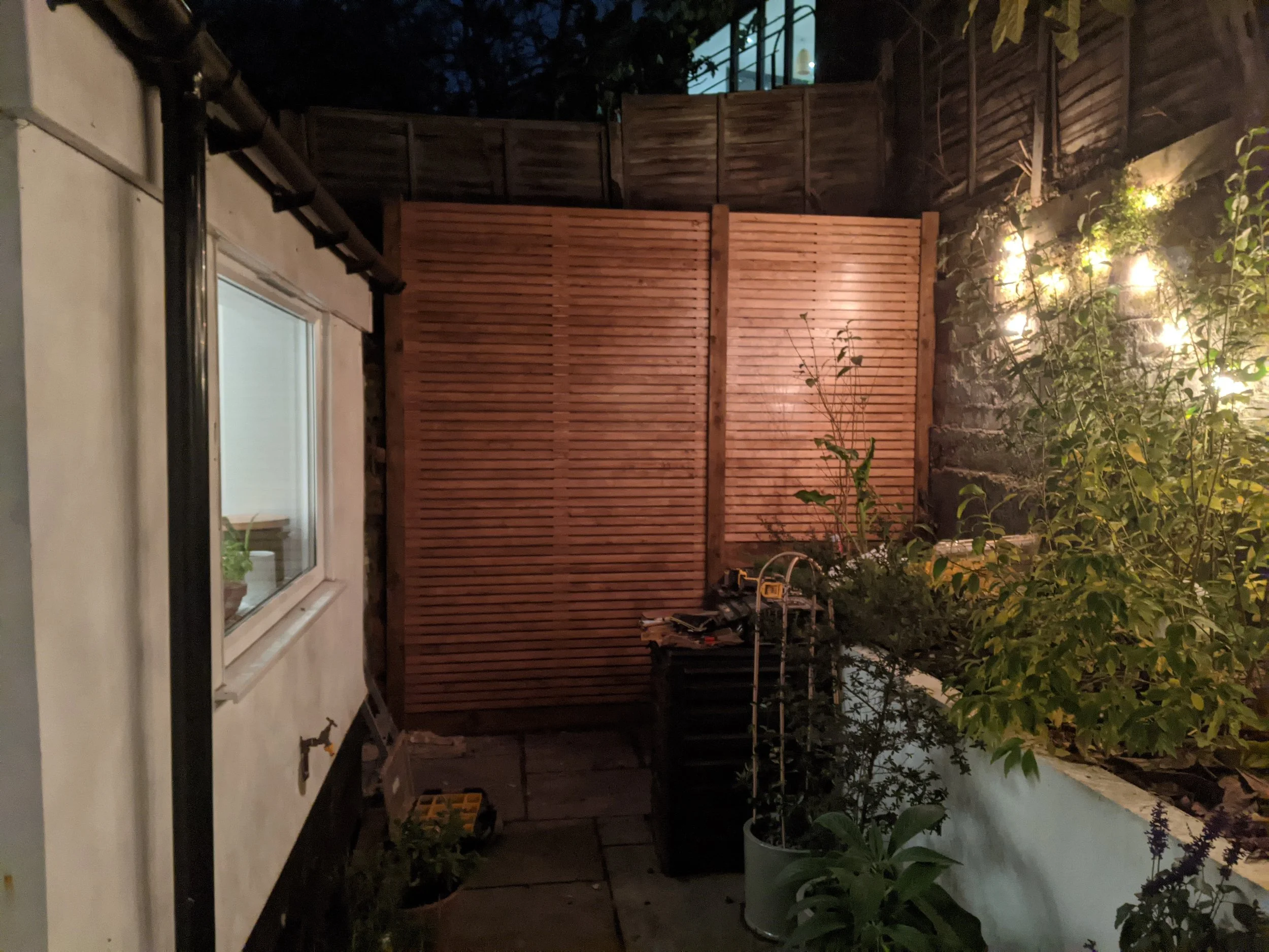 Nighttime backyard scene with a window on a white house, a wooden fence, and potted plants with garden lights illuminating the area.