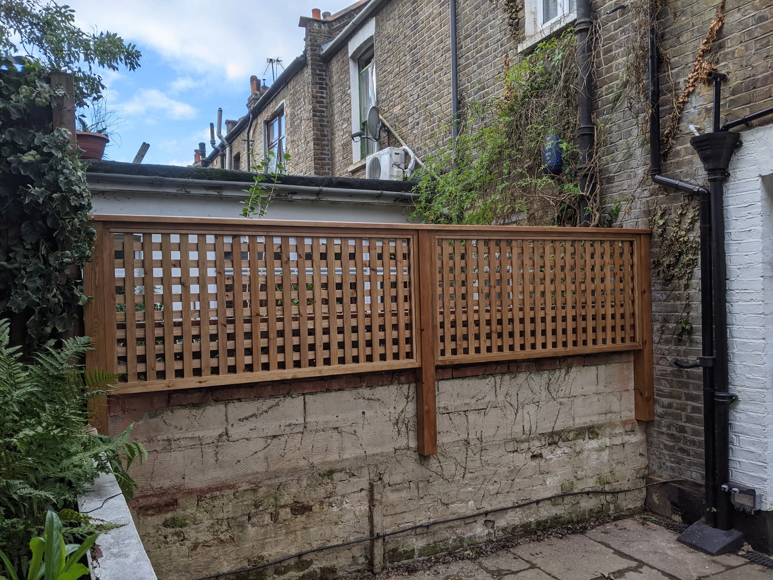 A back garden with a wooden fence on a brick wall, some greenery, and neighboring houses with brick exteriors and small windows.