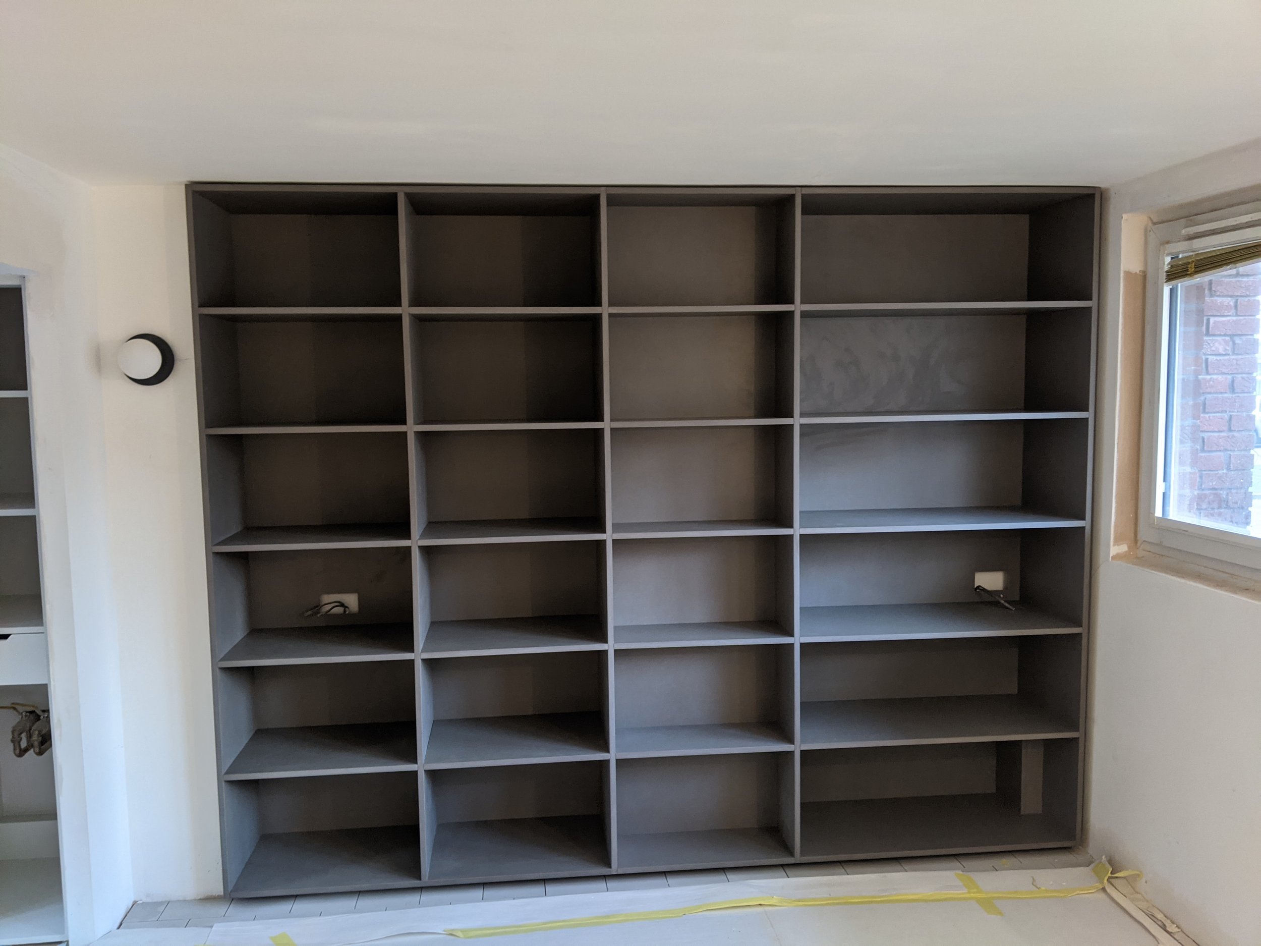 Empty gray bookshelf with multiple compartments in a room with a window on the right and a tiled floor.