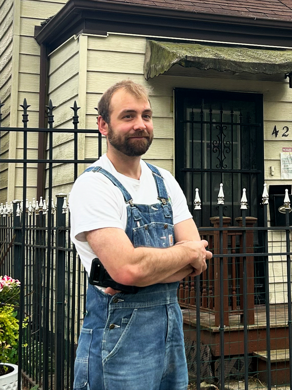 A man with a beard wearing a white t-shirt and denim overalls standing outside in front of a black metal fence and a house with beige siding, a black door with the number 42, and some potted plants.