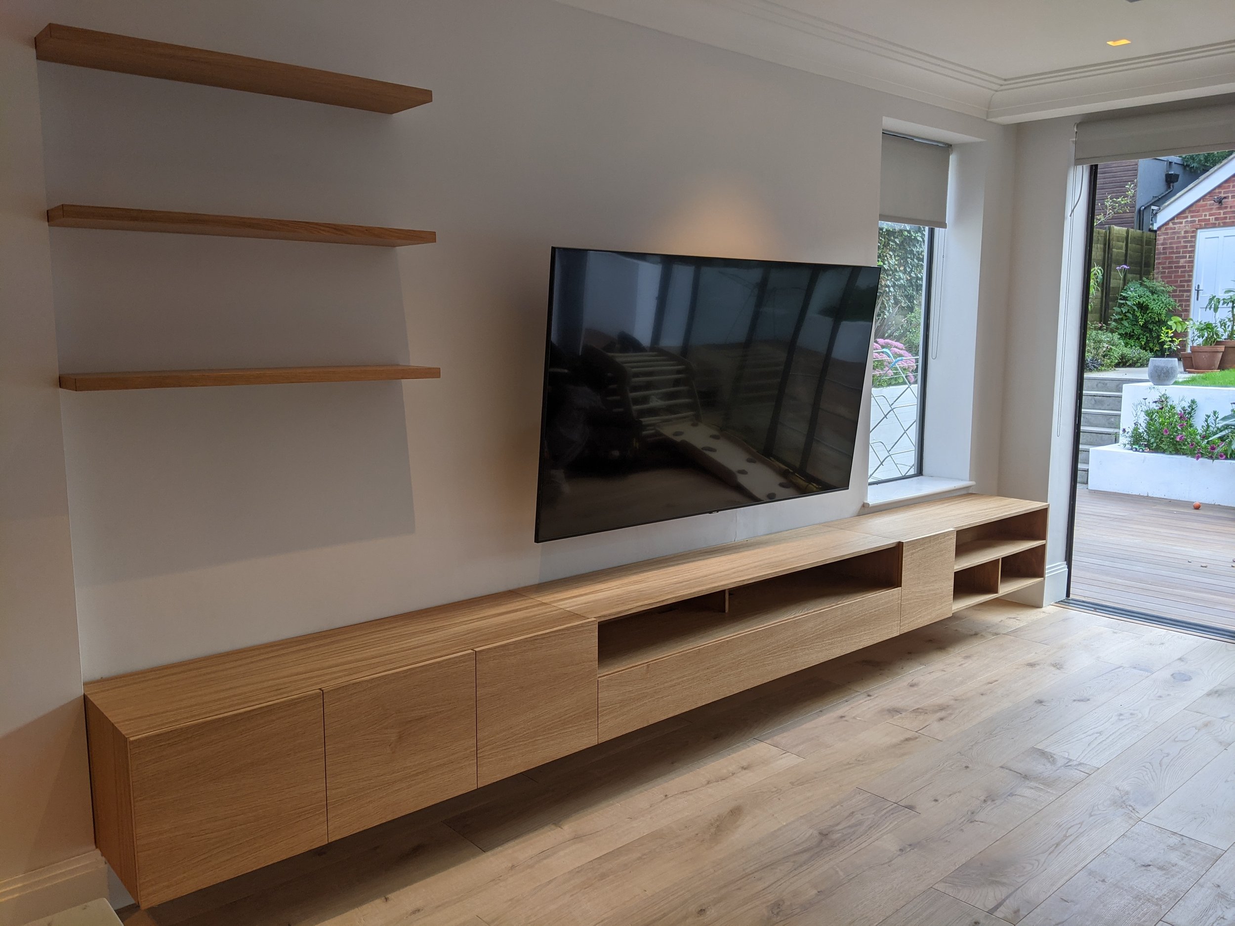 Modern wooden entertainment center with shelves and cabinets against a white wall, in a living room with large windows showing a garden outside.