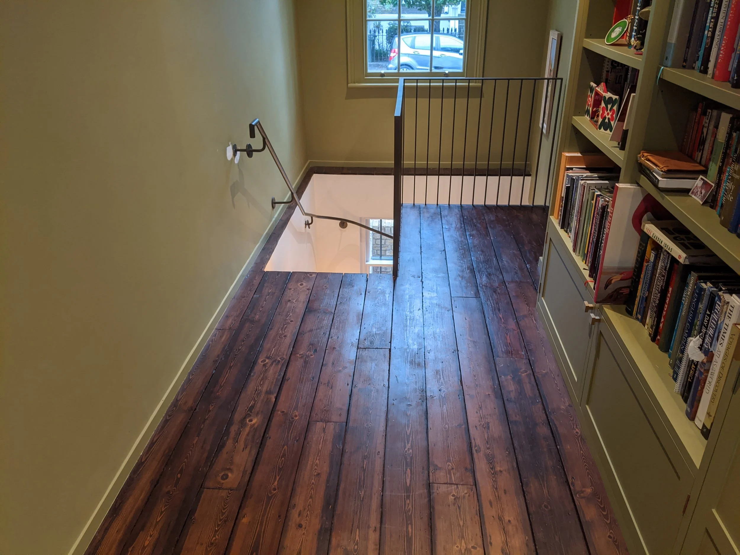 Interior of a house with dark wooden flooring, beige walls, a window with a car visible outside, a staircase, and a white bookshelf filled with books and decorations.
