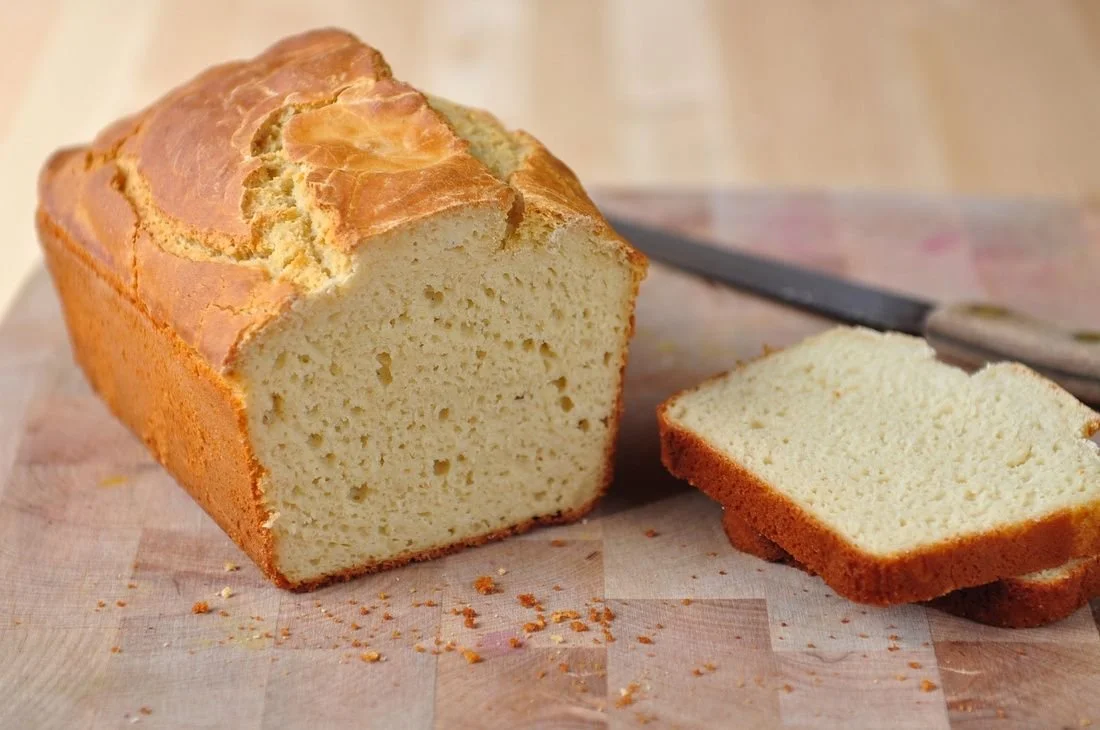 A loaf of rice bread with a golden brown crust and a light, fluffy interior on a wooden cutting board with a knife in the background.