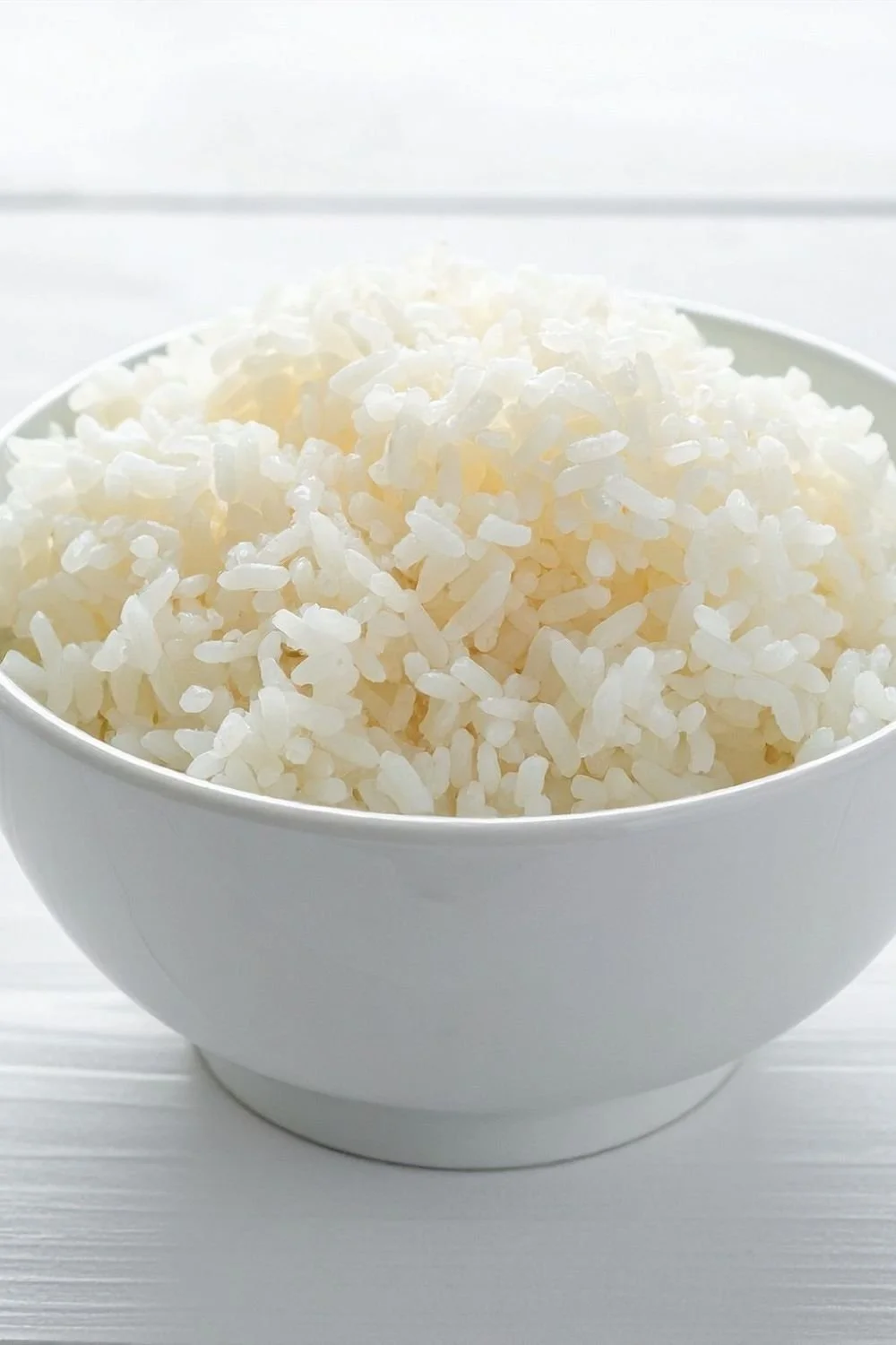 A white bowl filled with cooked white rice on a white surface with a white background.
