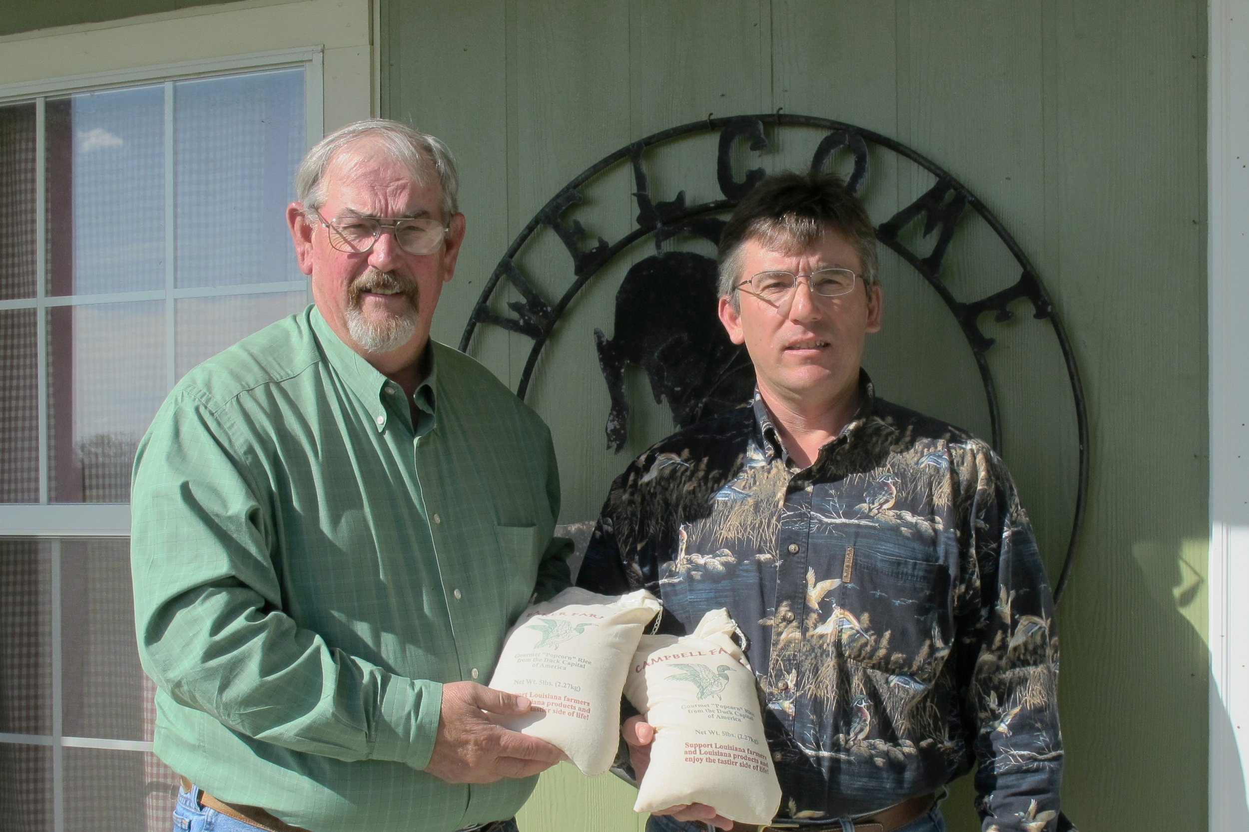 Roy Baker Sr. & Roy Baker Jr. standing outside in front of a green wall holding packages of rice from Louisiana.