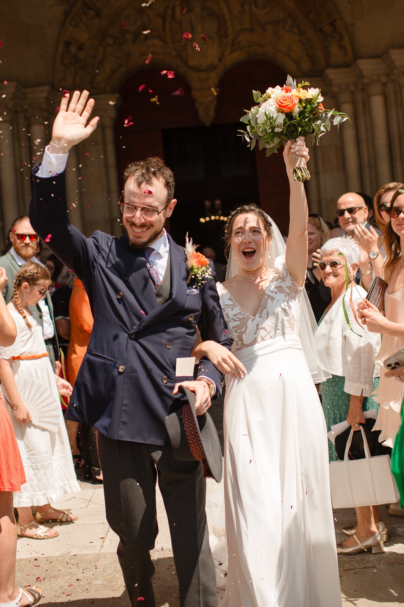 photographe mariage bordeaux photo de mariage wedding photo Un couple de mariés joyeux, la mariée en robe blanche, tenant un bouquet de fleurs, saluant la foule lors de leur mariage en plein air, entourés de convives souriants et applaudissant.