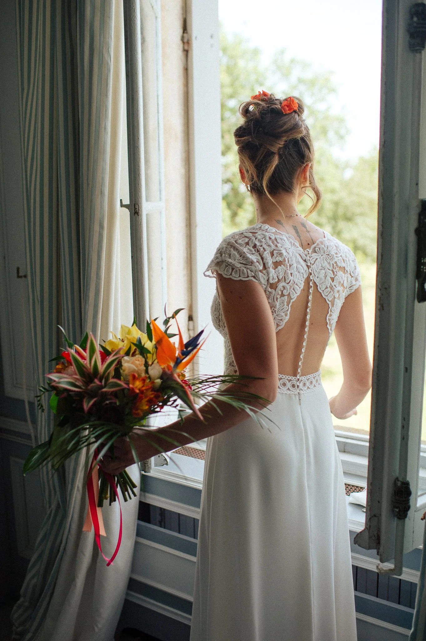 photographe mariage bordeaux photo de mariage wedding photo Une mariée en robe blanche regarde par une fenêtre, tenant un bouquet de fleurs colorées