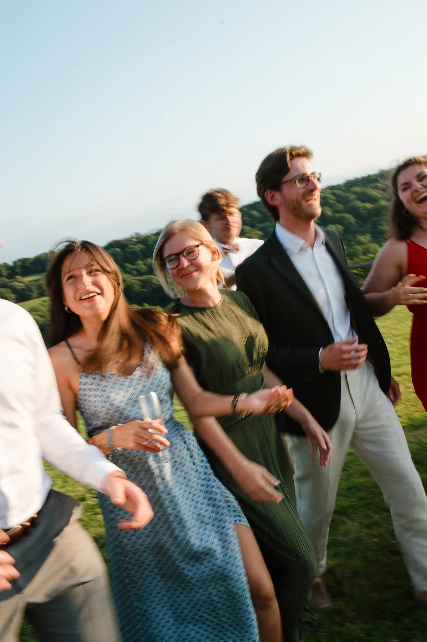 photographe mariage bordeaux photo de mariage wedding photo Groupe de personnes souriantes lors d'une fête en plein air, certains tenant des flûtes à champagne, avec un paysage verdoyant en arrière-plan.