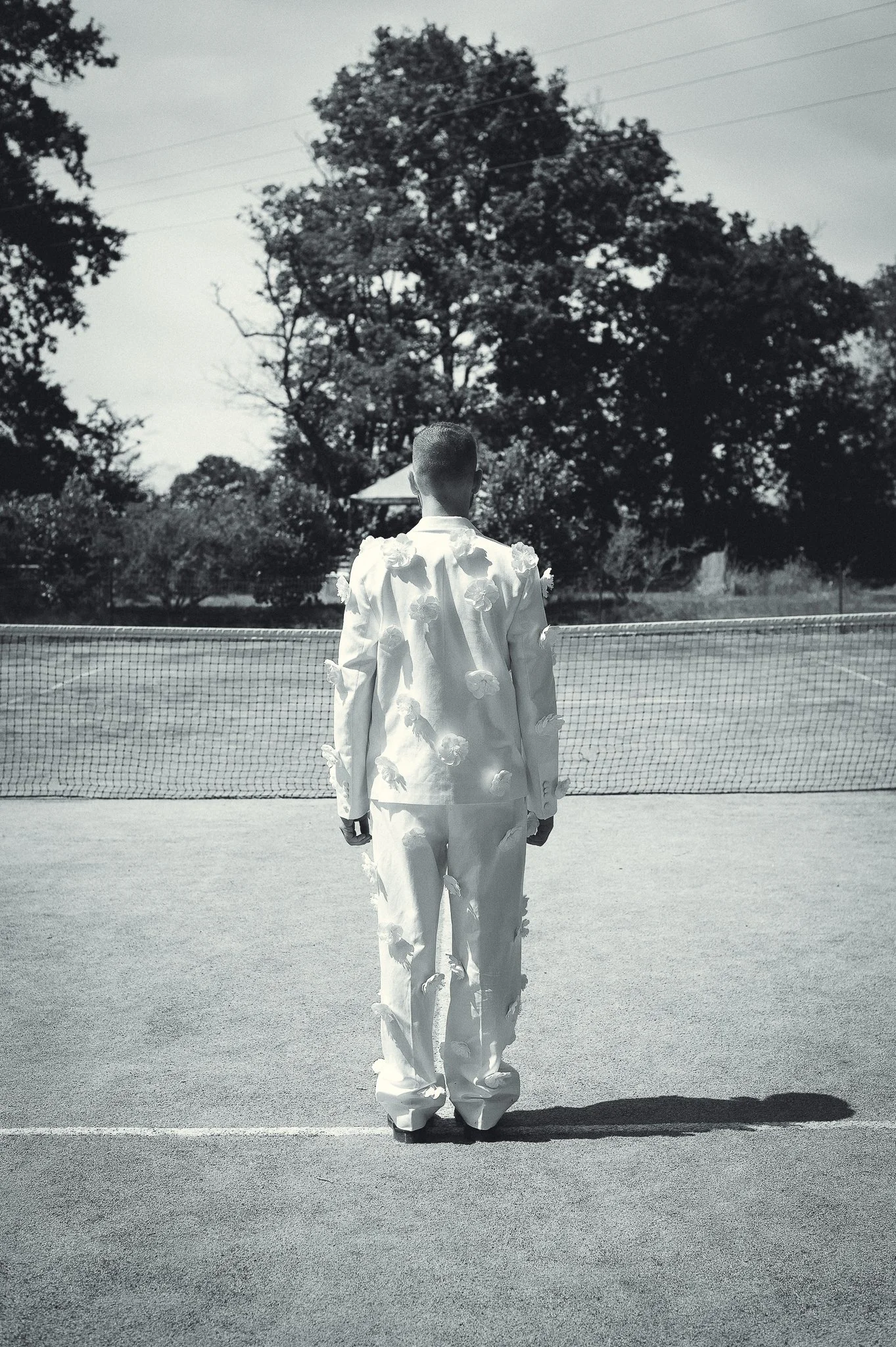 portrait mode fashion photo portrait homme photo bordeaux Une personne en costume blanc orné de fleurs, vue de dos, se tenant sur un court de tennis.