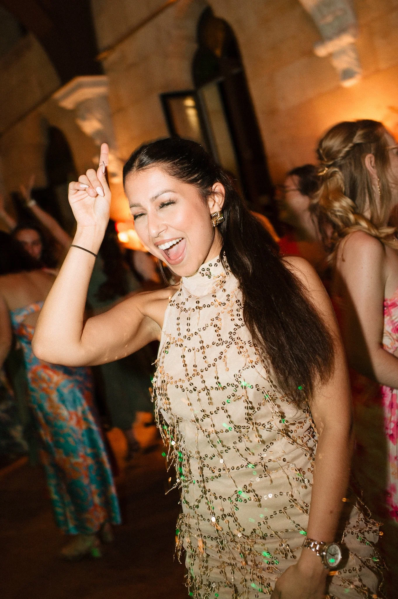 photographe événementiel bordeaux Une femme souriante et joyeuse qui danse lors d'une fête, portant une robe beige ornée de sequins dorés, dans une ambiance chaleureuse avec d'autres personnes en arrière-plan.