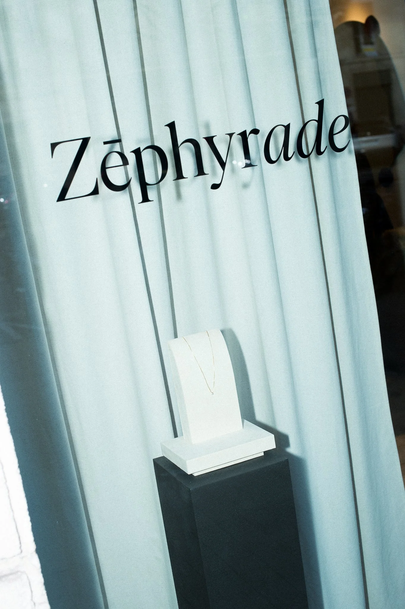 Vitrine de boutique avec le nom 'Zephygrade' écrit en noir, affichant un collier en or sur un support blanc, exposé sur un socle noir.