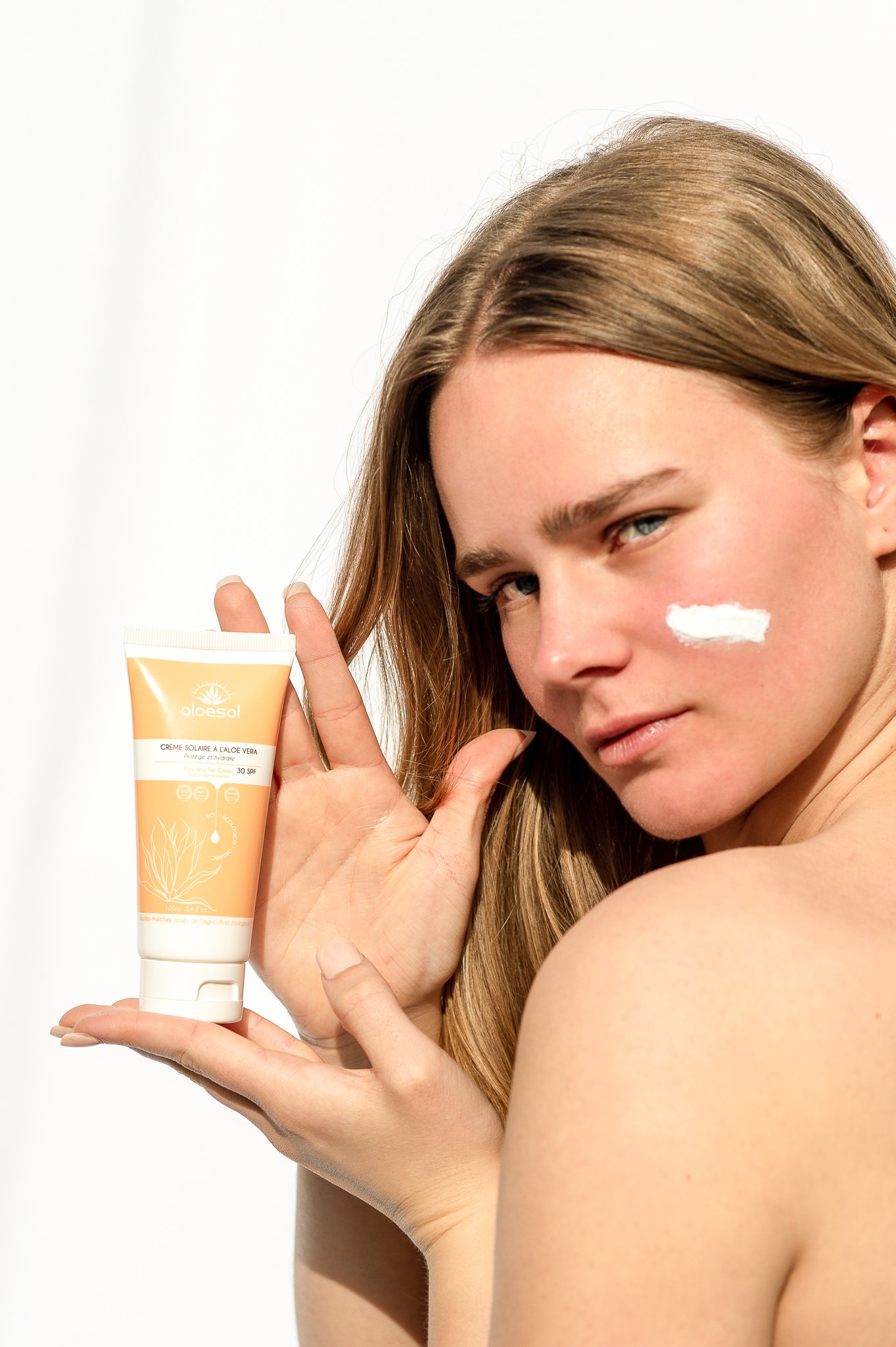 Une femme avec des cheveux longs et blonds tient un tube de crème solaire aloe avec SPF 30 près de son visage, avec une touche de crème sur sa joue.