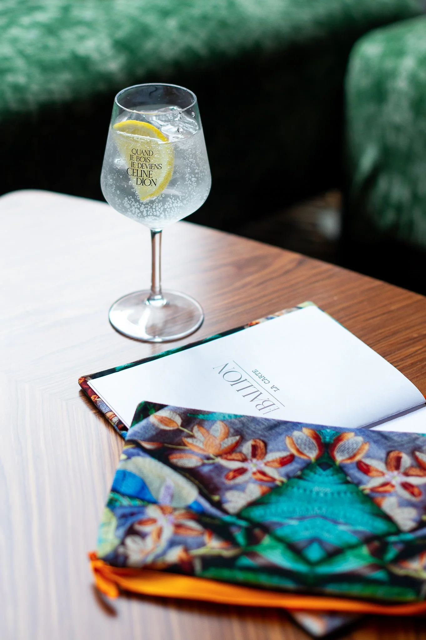 Verre d'eau gazeuse avec une tranche de citron sur une table en bois, un menu et un foulard coloré posé à côté.