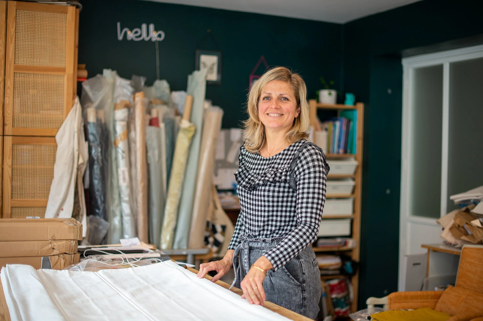 Femme souriante dans un atelier avec des rouleaux de tissu en arrière-plan et un bureau avec des piles de papiers.