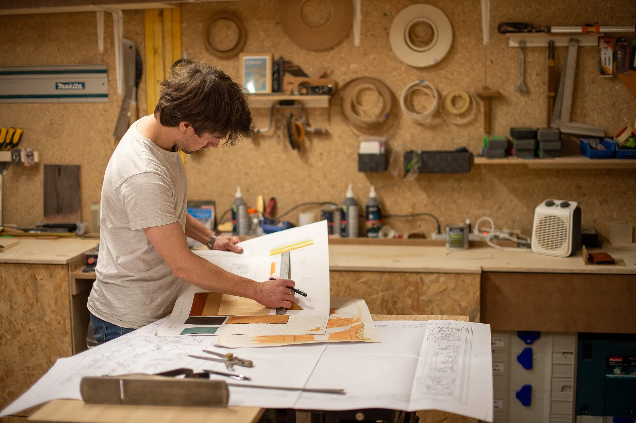 Un homme travaille dans un atelier de menuiserie, observant un plan ou un échantillon de matériau sur une grande table, avec des outils et matériaux variés autour.