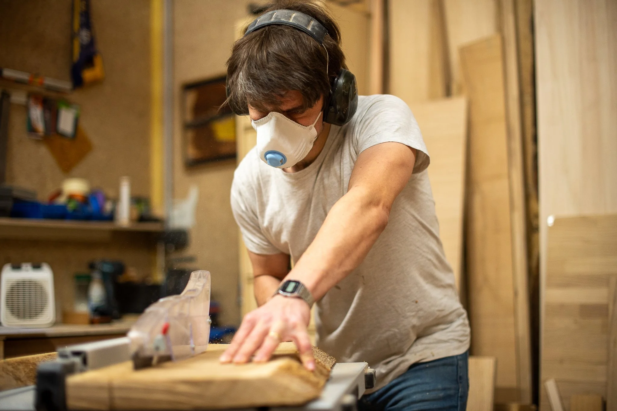 Homme portant un masque de protection et des bouchons d'oreilles, utilisant une scie pour couper du bois dans un atelier de menuiserie.