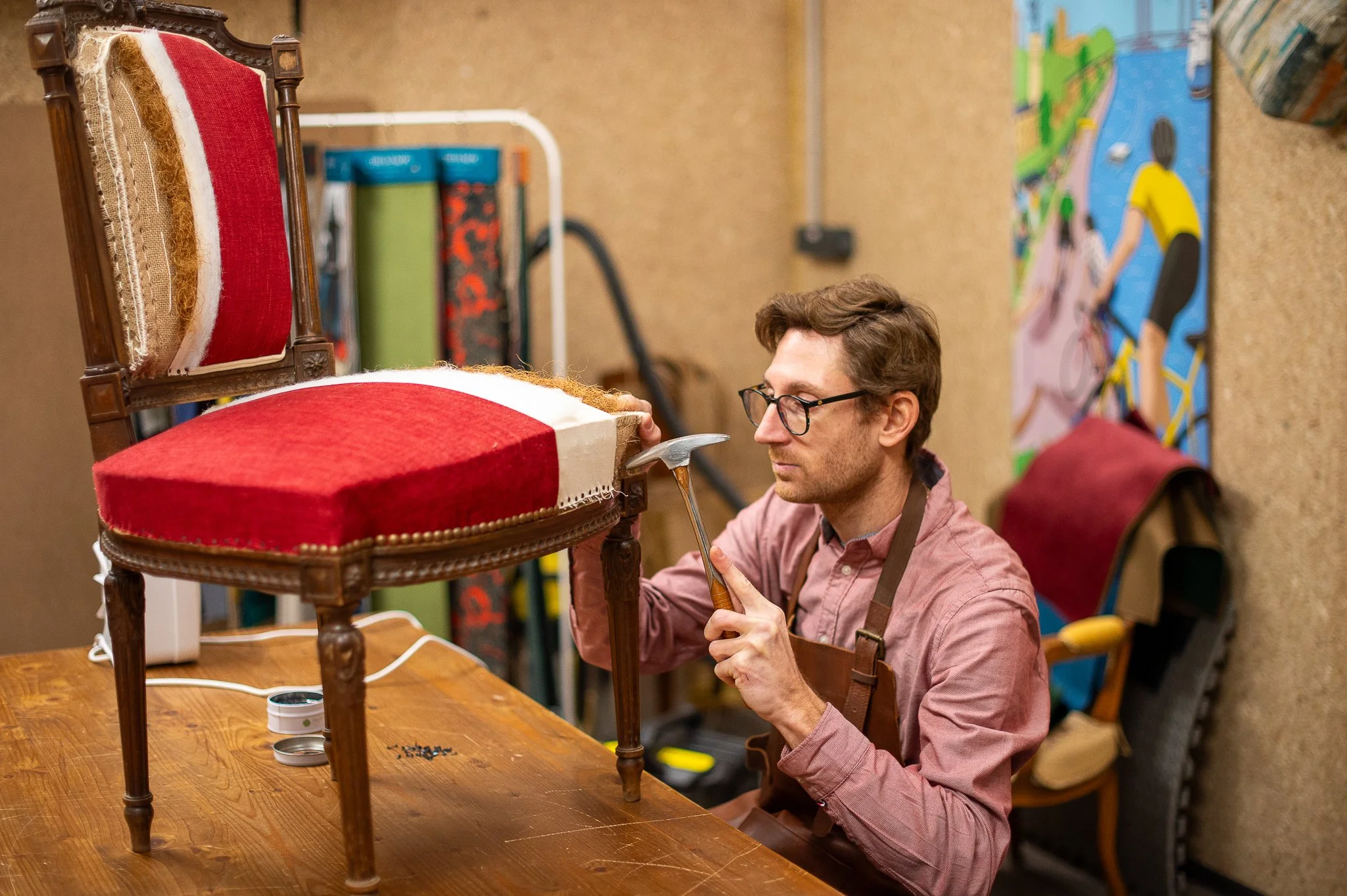 Un homme répare une chaise ancienne avec un marteau dans un atelier, à côté d'une peinture murale colorée.